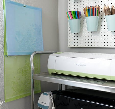 A peg board with markers in little buckets, a Cricut machine on a cart and Cricut mats hanging from a hook on the wall