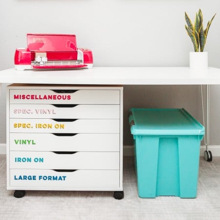 An aqua blue tote under a table that has a Cricut machine on top of it and a plant. The table has drawer's underneath that are labeled as "Miscellaneous", "Spec Vinyl", "Spec Iron On", "Vinyl" and "Iron On" and "Large Format"