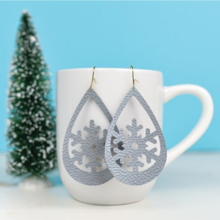 Close up of snowflake design earrings hanging from a coffee cup that is sitting on a table next to a small fake tree